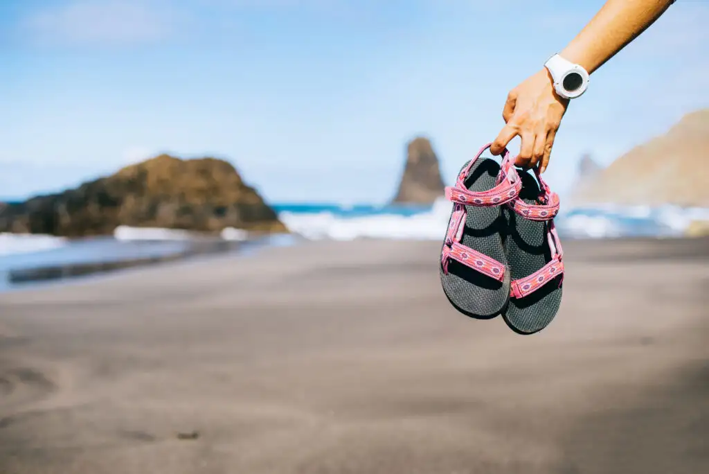 A hand holding a pair of sandals with the beach out of focus in the background