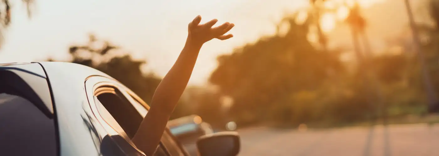 Person sticking hand out of window of car as car drives down street at sunset