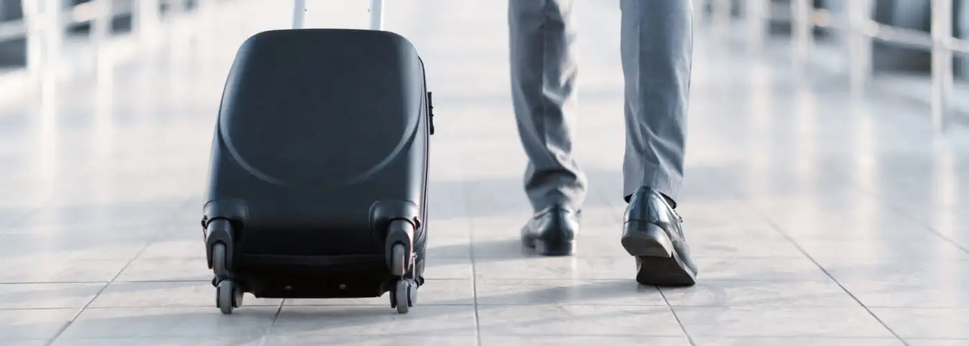 Man walking down a glass hallway in an airport pulling along a rolling carry-on suitcase