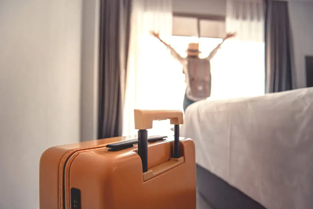 Person standing with arms in the air by a window with yellow suitcase in the foreground