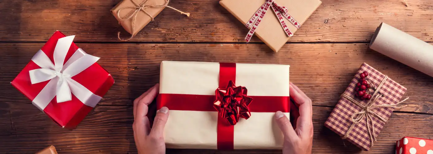 Aerial view of hands holding a gift surrounded by other affordable holiday travel gifts on a wooden table