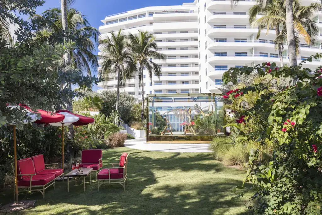 Looking at the rear of the Faena Hotel Miami Beach with palm trees, a table with chairs and umbrella, & bones of dinosaur on display