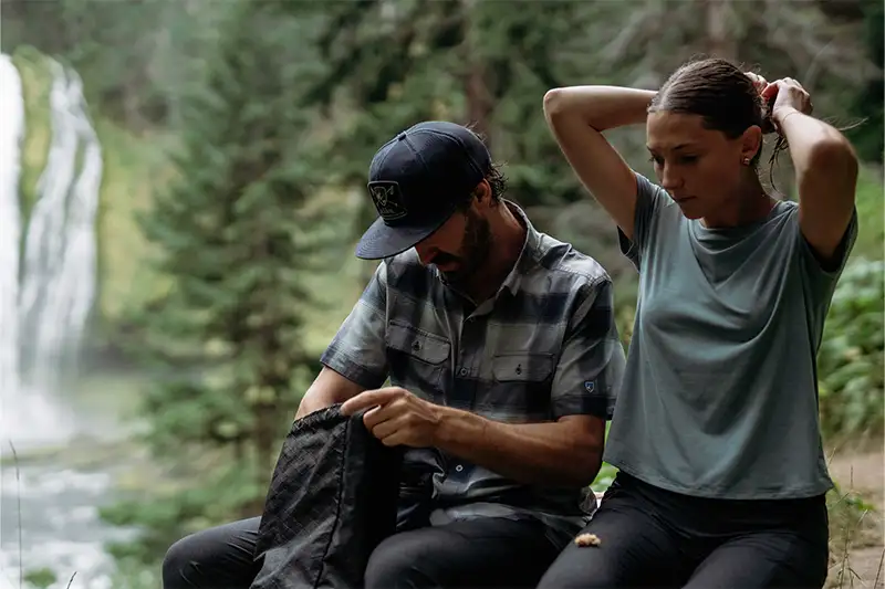 Couple sitting on a rock taking a break from hiking, the man is wearing a plaid button down and the woman is wearing Inspira Short Sleeve