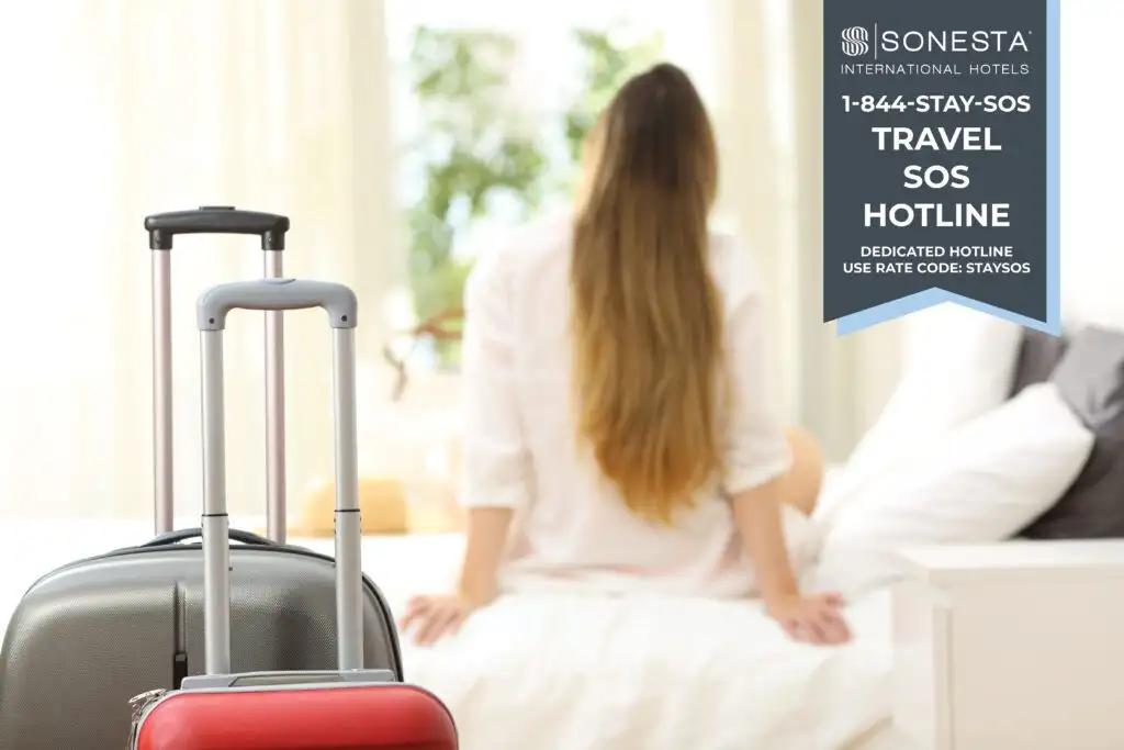 Woman sitting on a hotel bed facing away from the camera with two rolling suitcases in the foreground